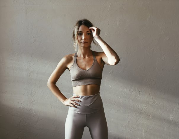 Woman in workout clothes stands in a gray room while brushing her hair back after CoolSculpting and Emsculpt NEO treatment